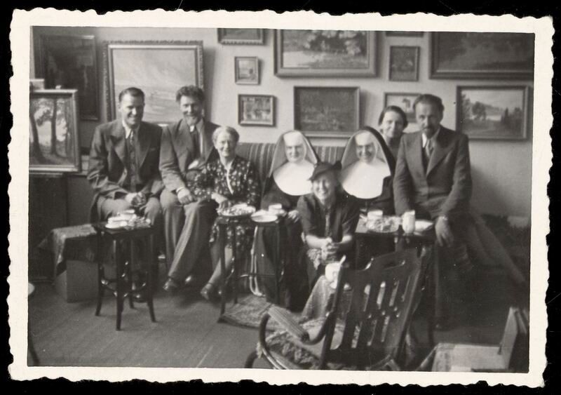 Dirbtuvėje V. Putvinskio g. 40 ant sofos prie kilnojamųjų mažų staliukų su kavos puodeliais. Pirmas iš kairės krepšininkas P. Lubinas, šalia A. Žmuidzinavičius, M. Žmuidzinavičienė, dvi vienuolės, M. ir J. Nemeikšos ir nežinoma moteris su skrybėle. 1939. ČDM ŽFp 271.