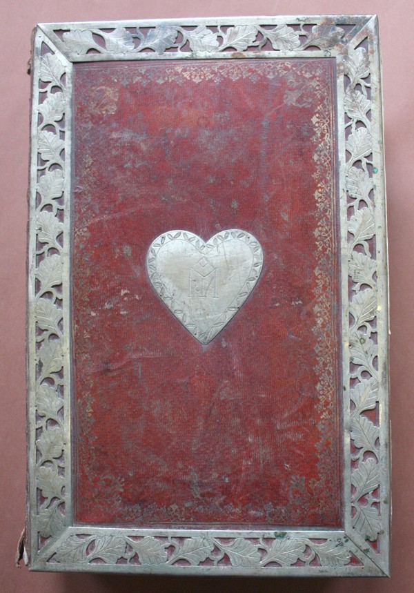 Metaliniais apkaustais su lapų girliandos ornamentu įrėmintas kietviršis, kurio viduryje - metalinė širdies formos plokštele su išraižyta Marijos monograma.